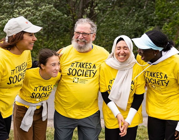 A group of people wearing ‘It takes a society” T-shirts are standing in front of a wooded area. They are laughing and smiling.